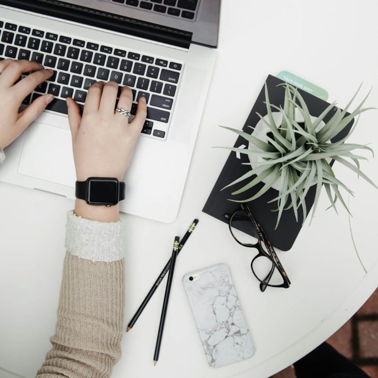 Hände tippen auf Laptop, daneben Pflanze, Notizbuch, Smartphone und Brille.